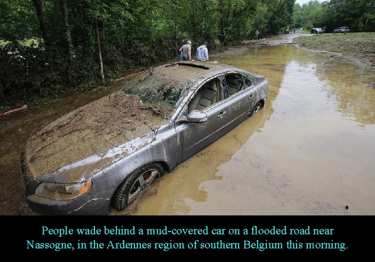People wade behind a mud-covered car on a flooded road near Nassogne, in the
