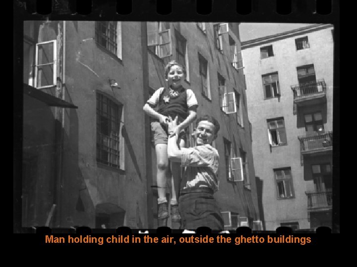 Man holding child in the air, outside the ghetto buildings 