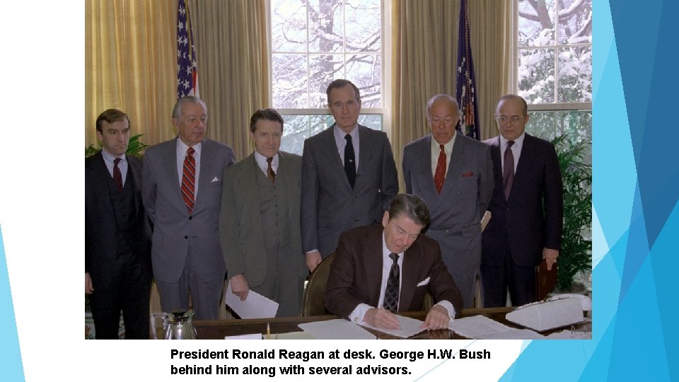 President Ronald Reagan at desk. George H. W. Bush behind him along with several